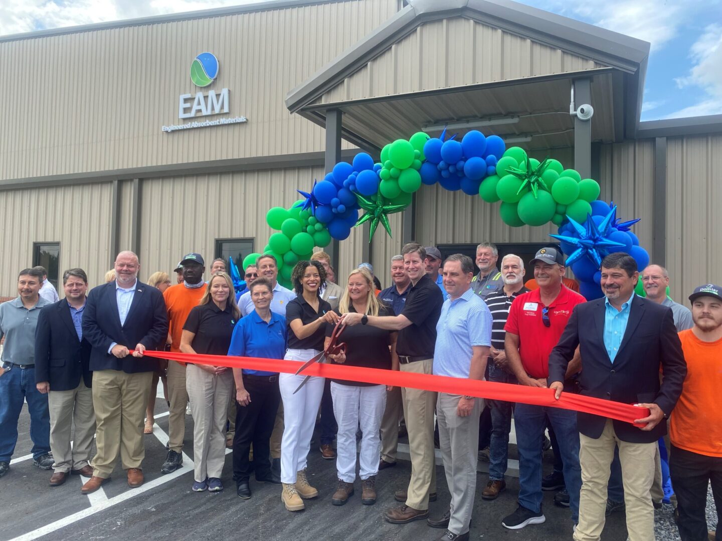 Domtar celebrated the EAM expansion in Jesup, GA on May 24. Pictured: Group of people in front of green and blue balloon arch and EAM facility exterior, with 3 people in the middle holding scissors to cut the ribbon on the new expansion. The ribbon is red-orange and being held by two men about 12 feet apart.
