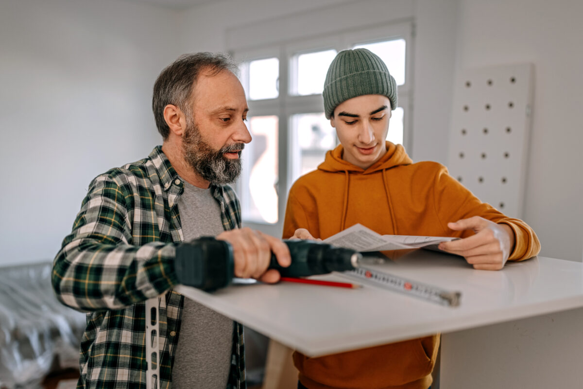 Senior man with teenage boy reading instructional manual while building DIY furniture and shelf during apartment renovation