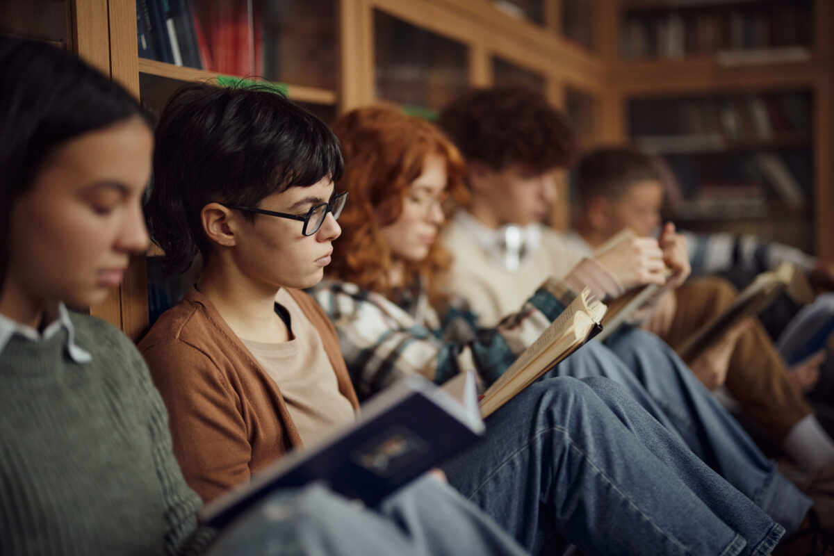 Group of high school students studying from books while relaxing on floor in library. Focus is on woman with eyeglasses.