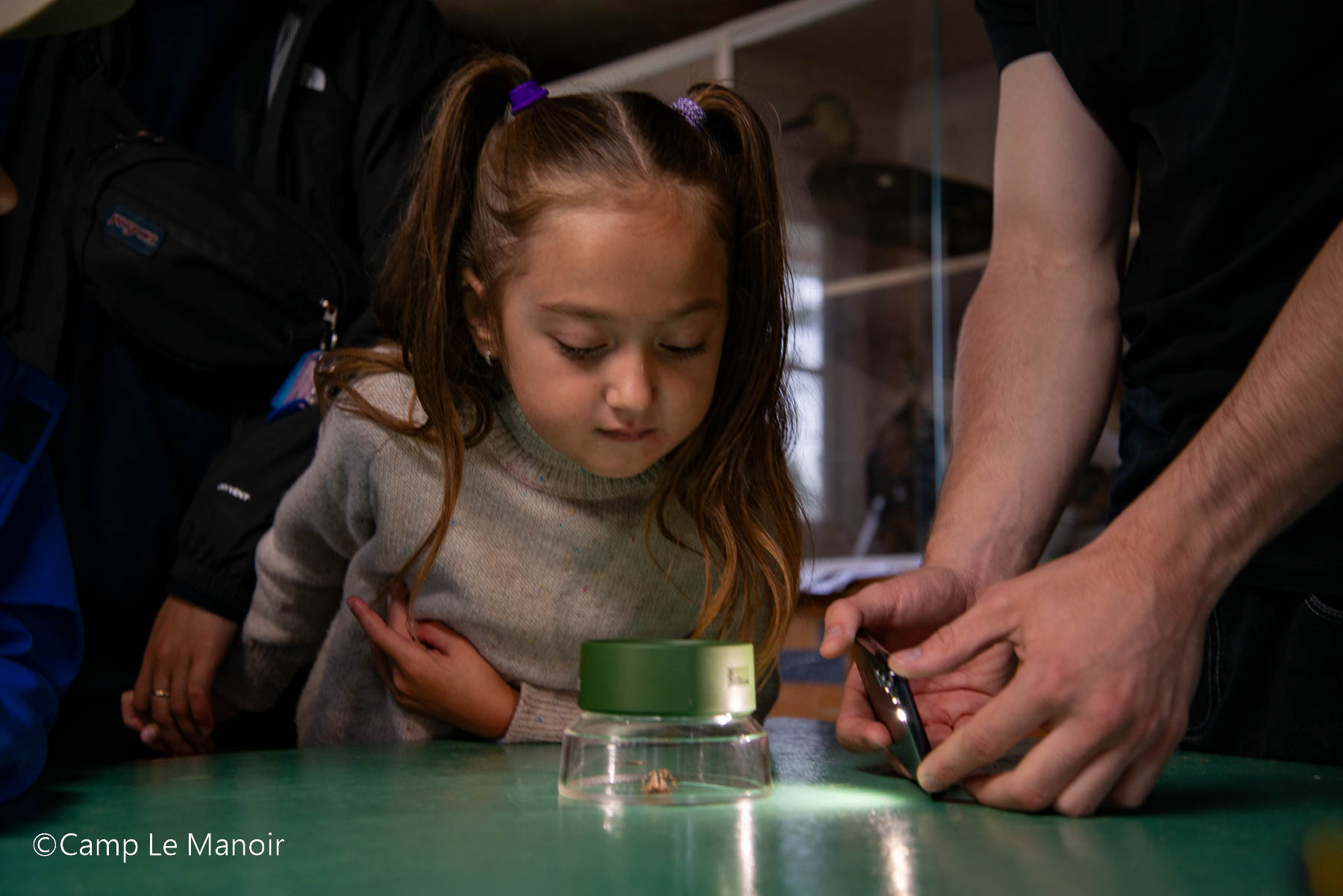 $2K Your Way grant supports Camp le Manoir in Quebec. Image description: Young girl looking at bug under light and magnifying glass with camp counselors nearby. Copyright Camp le Manoir