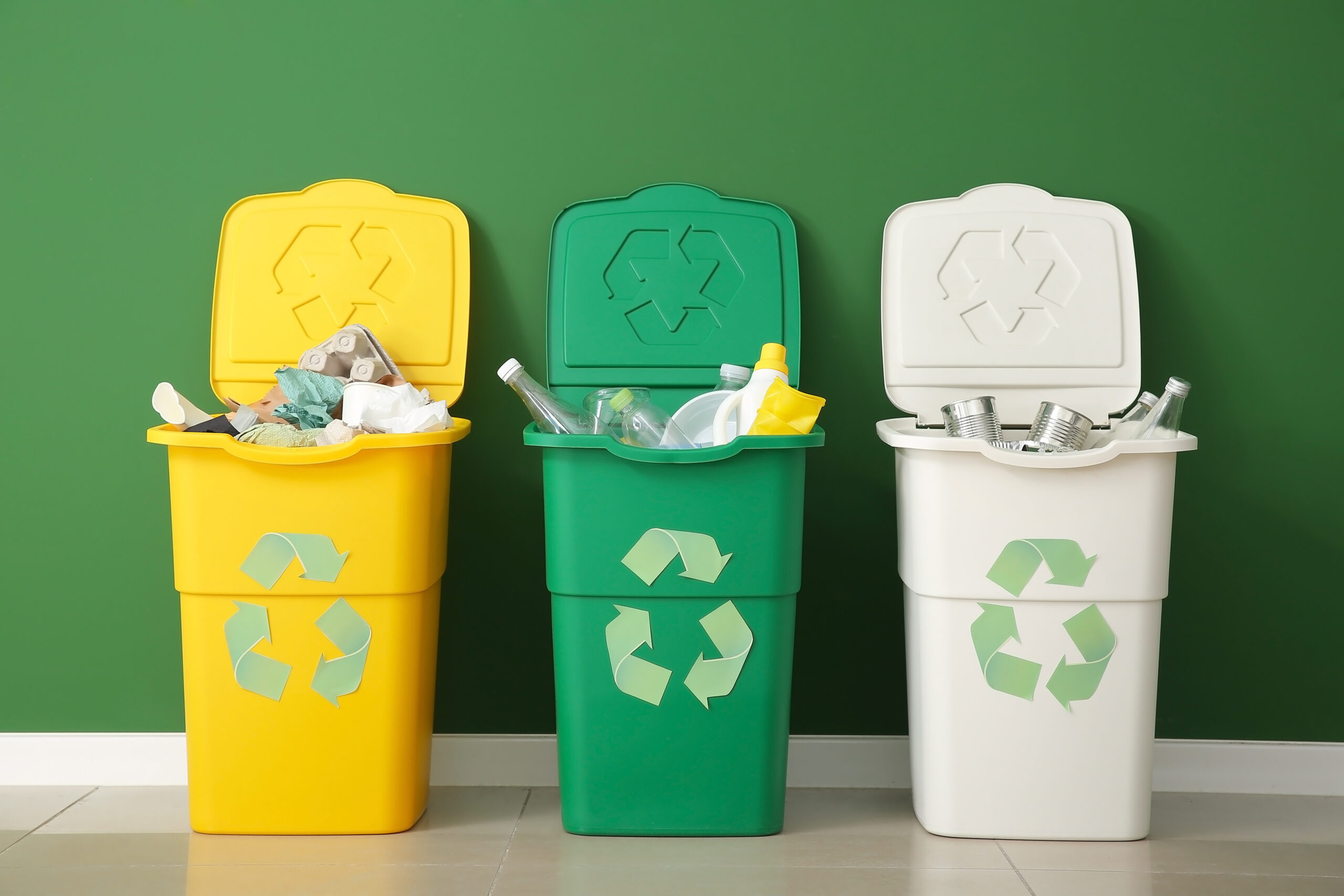 One yellow, one green and one white bin, all with the recycling symbol on them, filled with recyclables. They are up against a green wall. Check out Domtar's modern guide to curbside recycling.