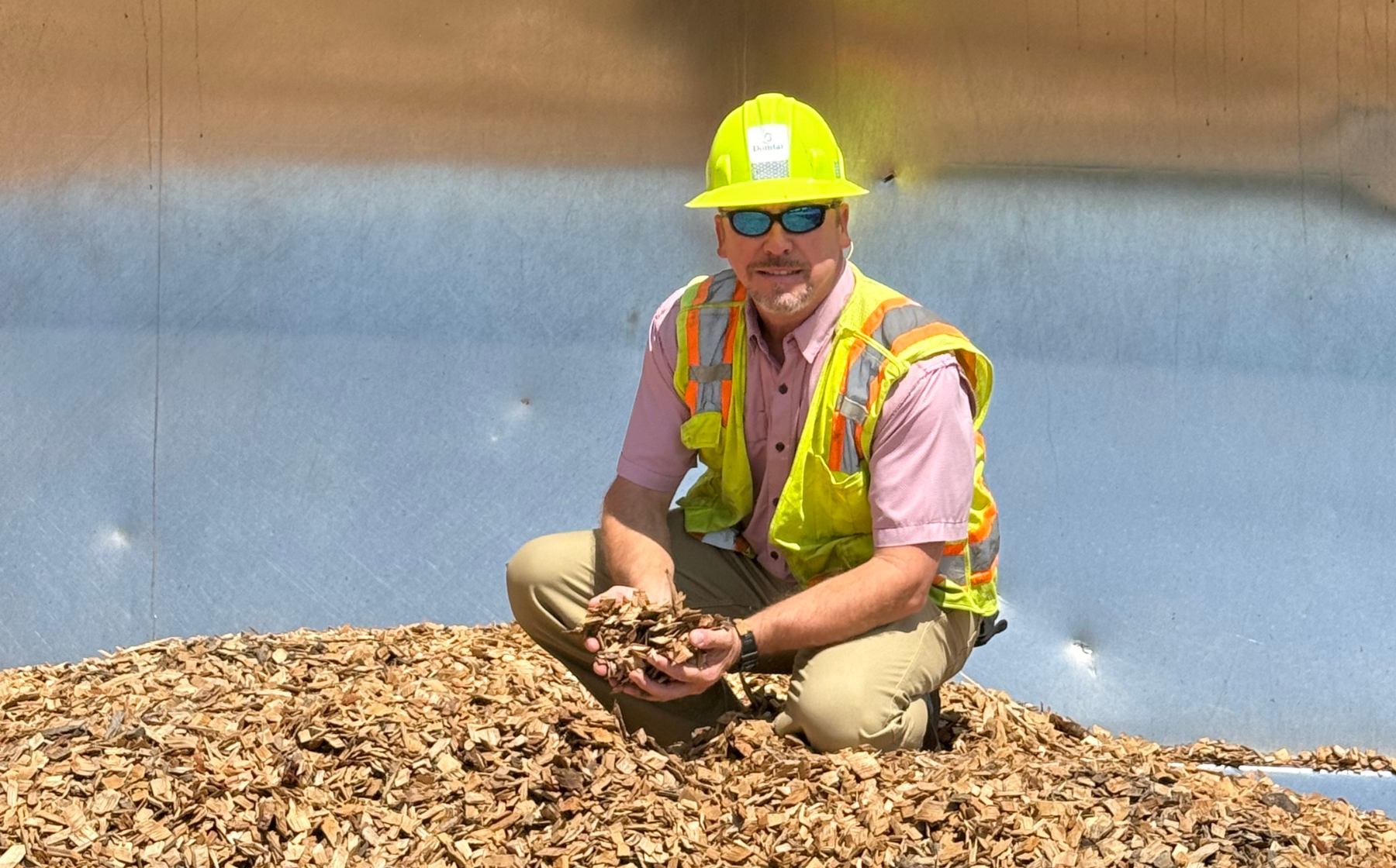 A man, Chad Robertson, crouched on top of a pile of wood chips, holding wood chips in his hand. He is wearing a yellow hard hat and safety vest over his work clothes. Chad leads a forest biodiversity partnership Domtar has developed with NatureServe in Mississippi.