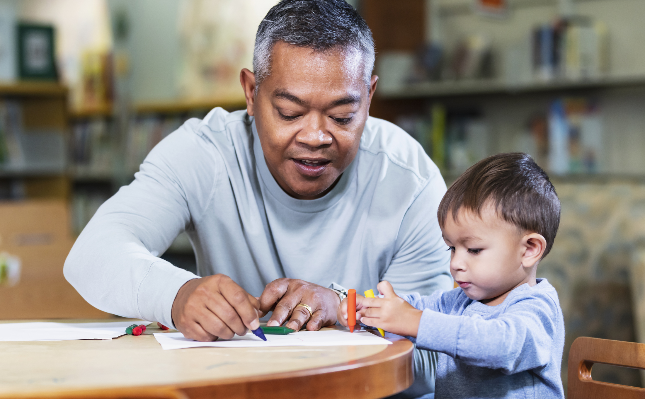 A father spending time with his son, a 19 month old toddler. They are sitting at a table coloring with crayons.